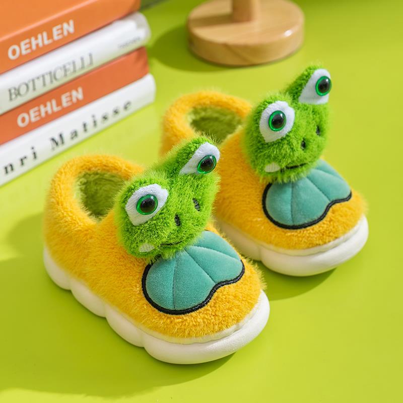 Green frog-themed children's yellow slippers on a green surface with books in the background.