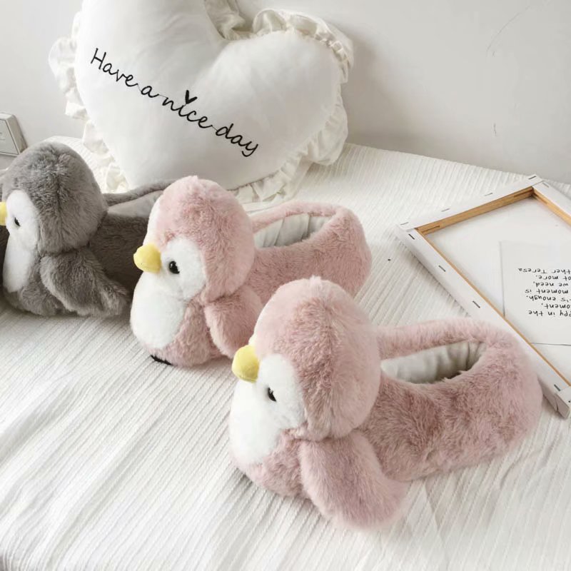 Pink penguin slippers on a light wooden floor with a heart-shaped pillow in the background.