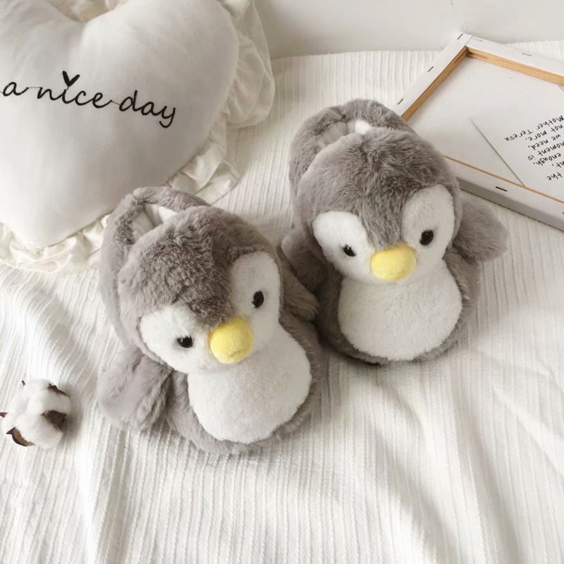 Pair of plush penguin slippers on a soft surface with a heart-shaped pillow in the background.