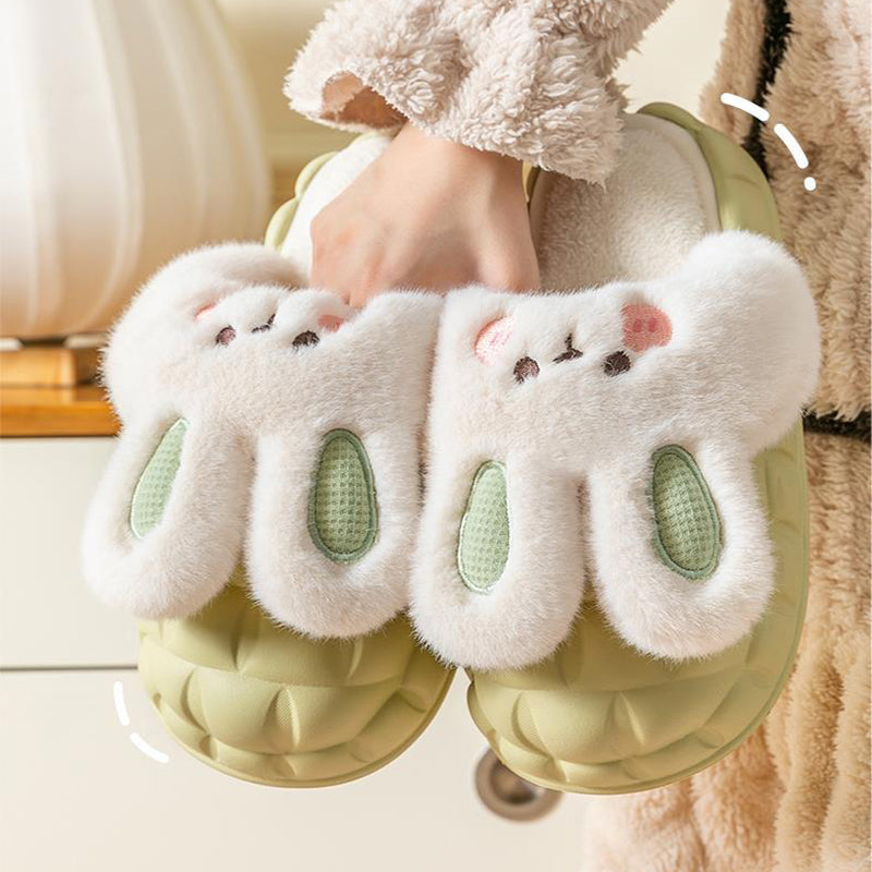 White fluffy rabbit slippers with green accents worn by a person, on a light background.