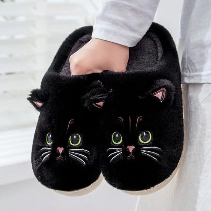Black cat-shaped slippers with green eyes and pink noses held by a person.