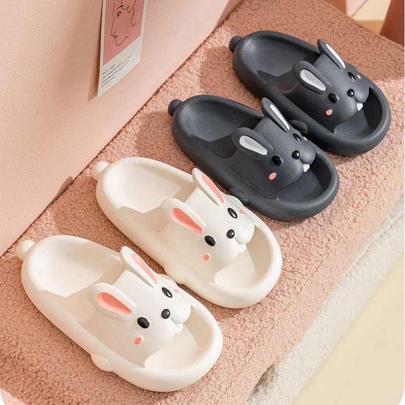 Pair of dark gray slippers with bunny face design held by hands against a pink background