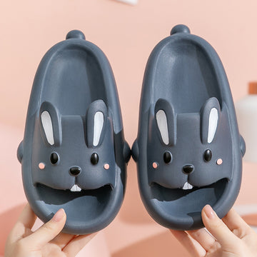 Pair of dark gray slippers with bunny face design held by hands against a pink background