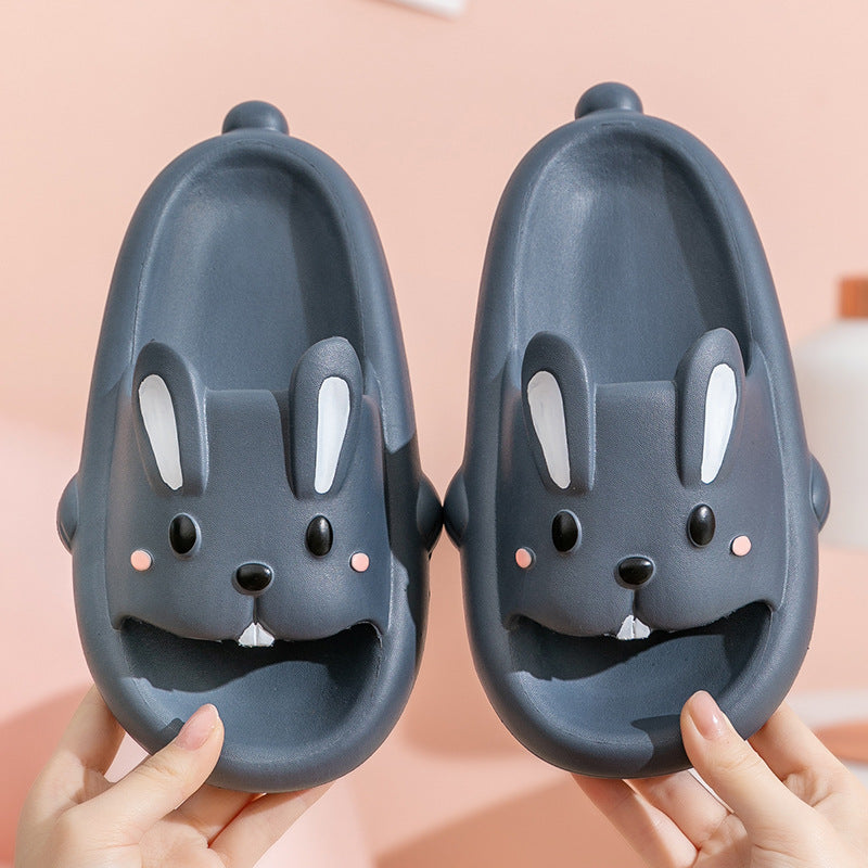 Pair of dark gray slippers with bunny face design held by hands against a pink background