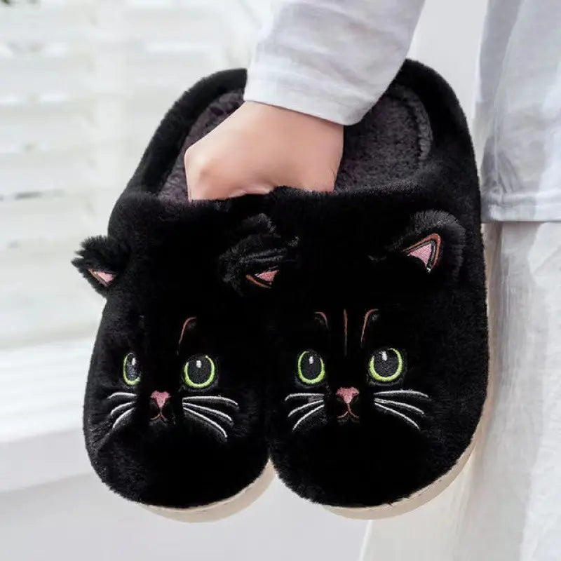 Black cat-shaped slippers with green eyes and pink noses held by a person.