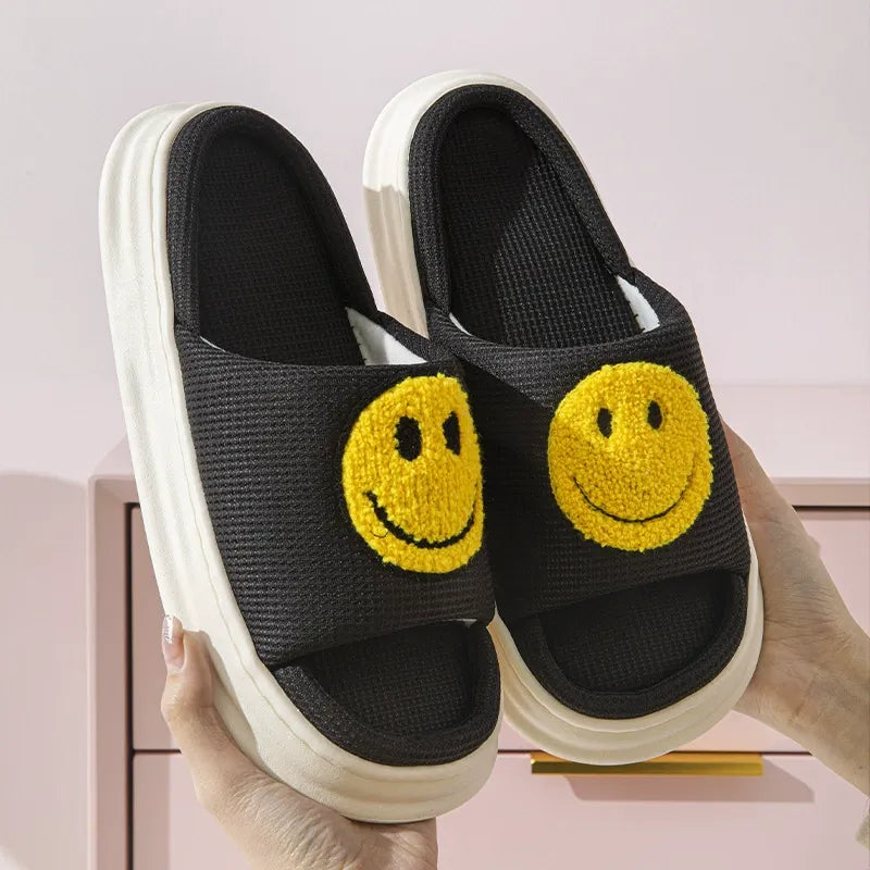 Black slip-on shoes with yellow smiley face designs held by a hand against a light pink background.