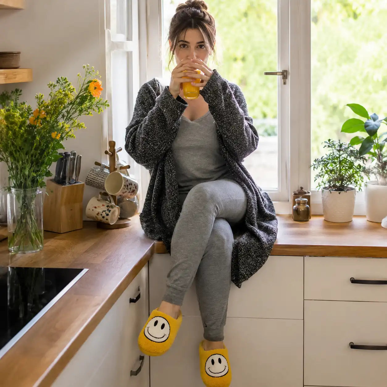 Woman sitting on a kitchen counter drinking from a glass, wearing yellow smiley face slippers.