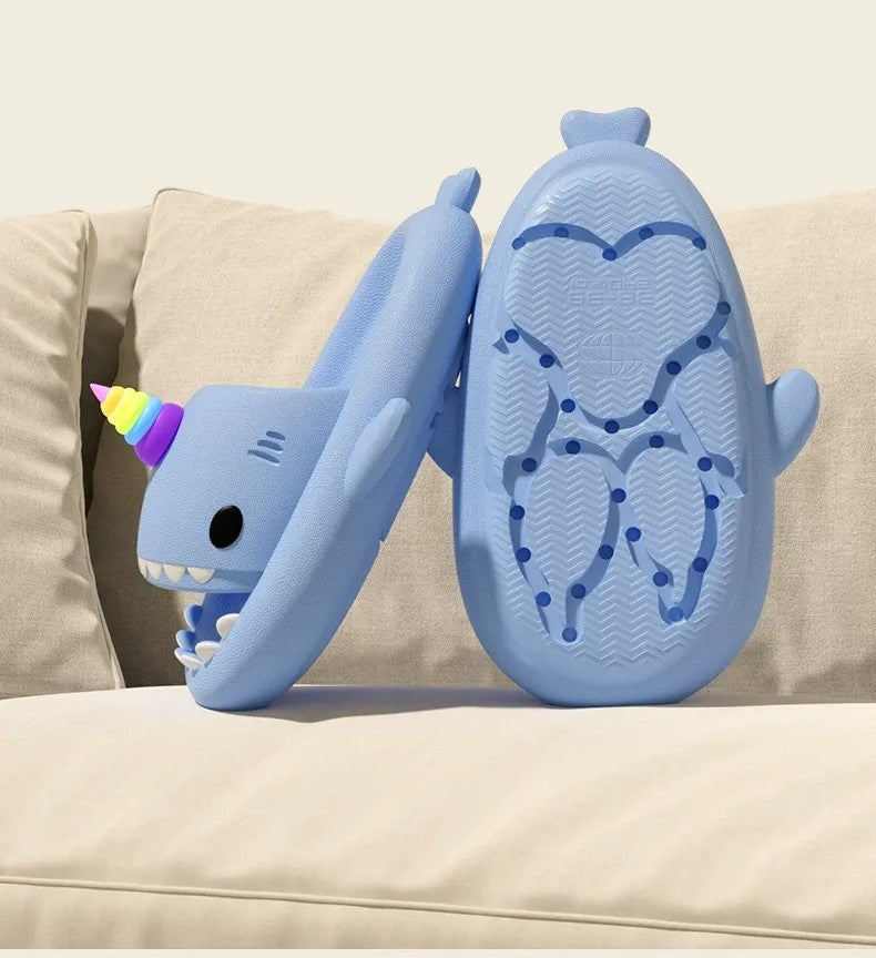 Blue shark-shaped slippers with a unicorn horn on a beige couch.