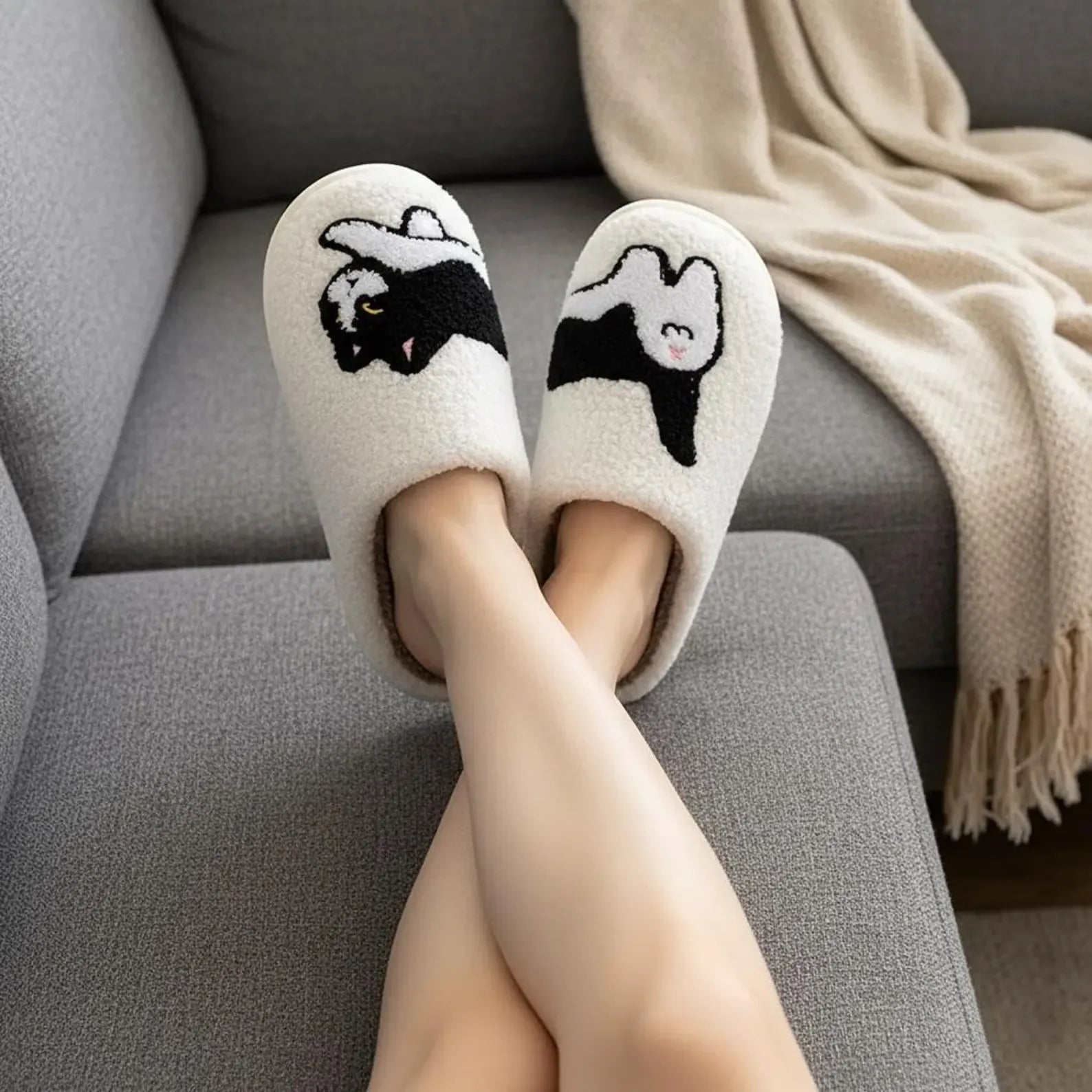 Person wearing white slippers with black cat designs on a gray sofa.