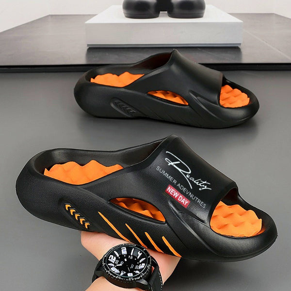 Black slides with orange sole held by a hand wearing a black watch on a gray surface.