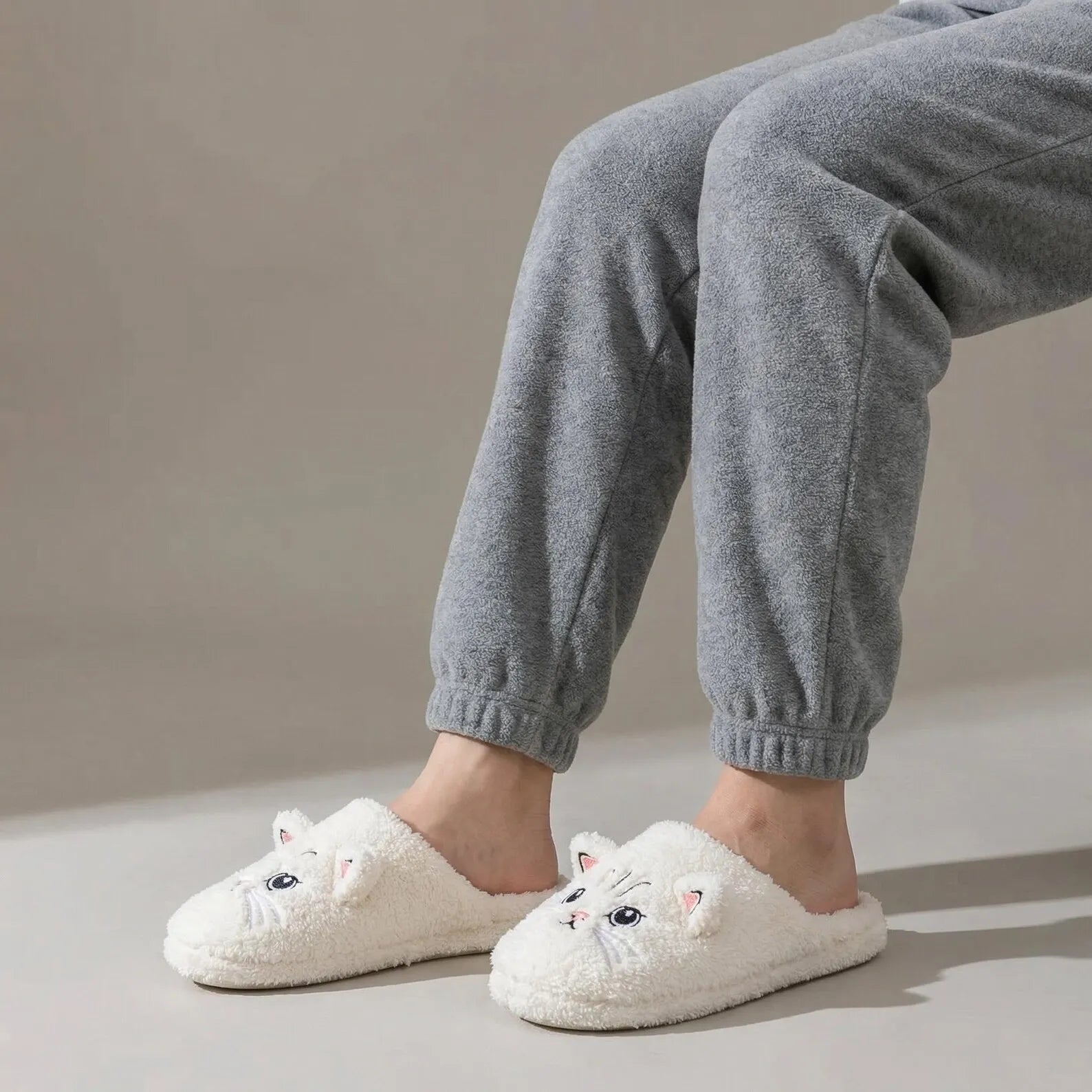 White fluffy slippers with cat face design worn by a person in gray pants on a neutral background