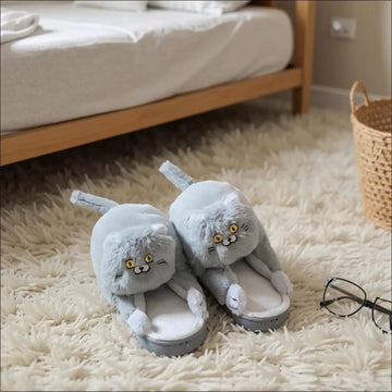 Gray slippers with cat face design on a fluffy carpet next to a bed.