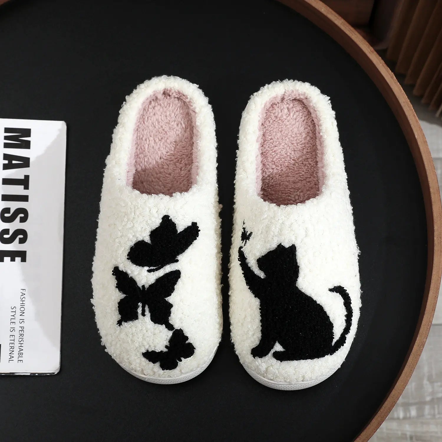 White slippers with black cat and flower design on a dark surface, featuring a Matisse card.