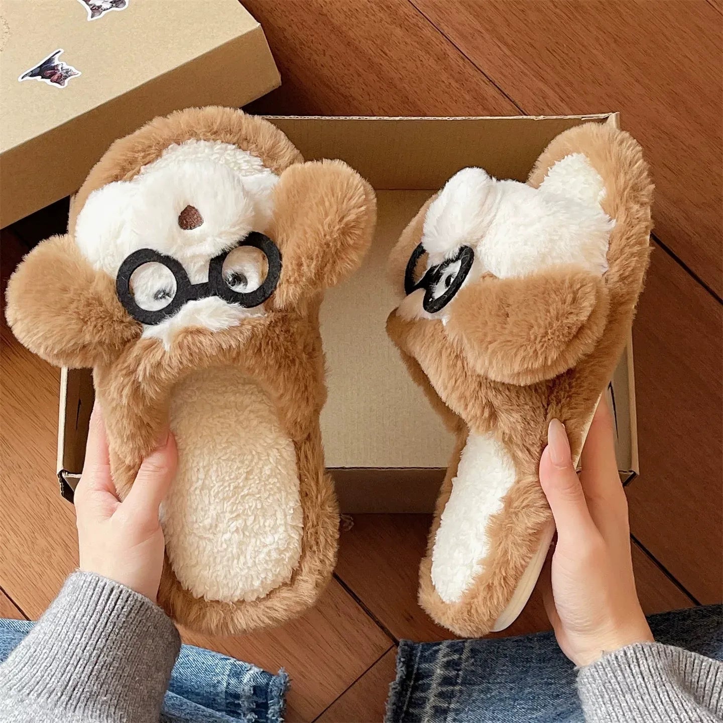 Pair of plush slippers with a monkey design and glasses held by two hands on a wooden floor.